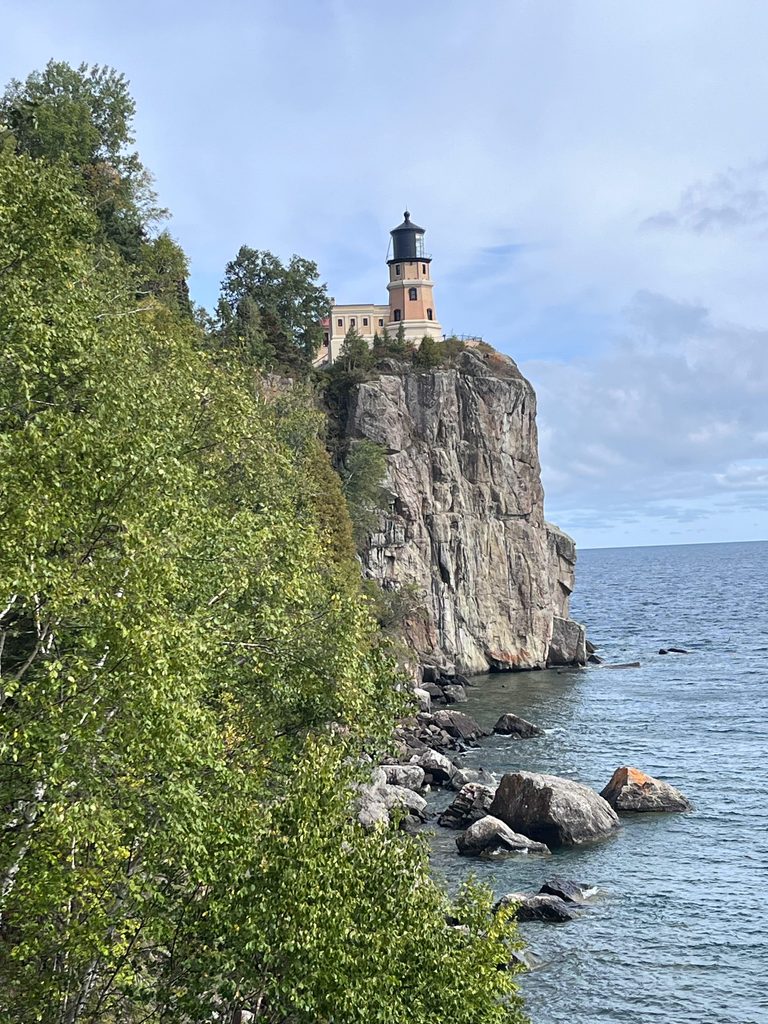 A lighthouse standing on a tall rocky cliff overlooking blue water, surrounded by green trees.