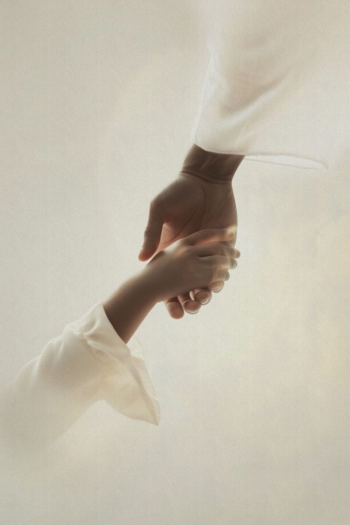 Two hands reaching toward each other, one gently holding the other against a soft cream background.