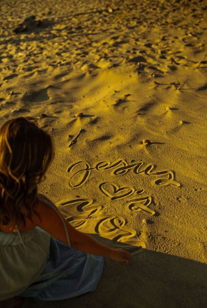 A woman sitting on a beach beside the words Jesus Loves You written in the sand.