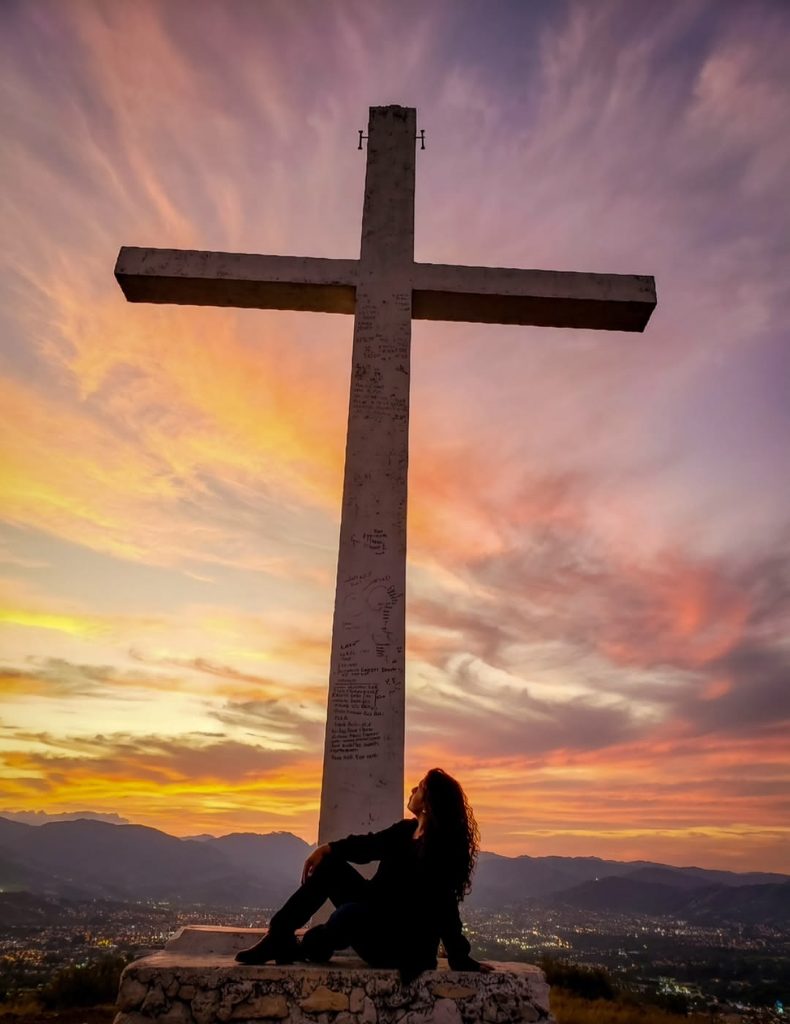 A woman sitting beneath a large cross on a hill overlooking the mountains at sunset.