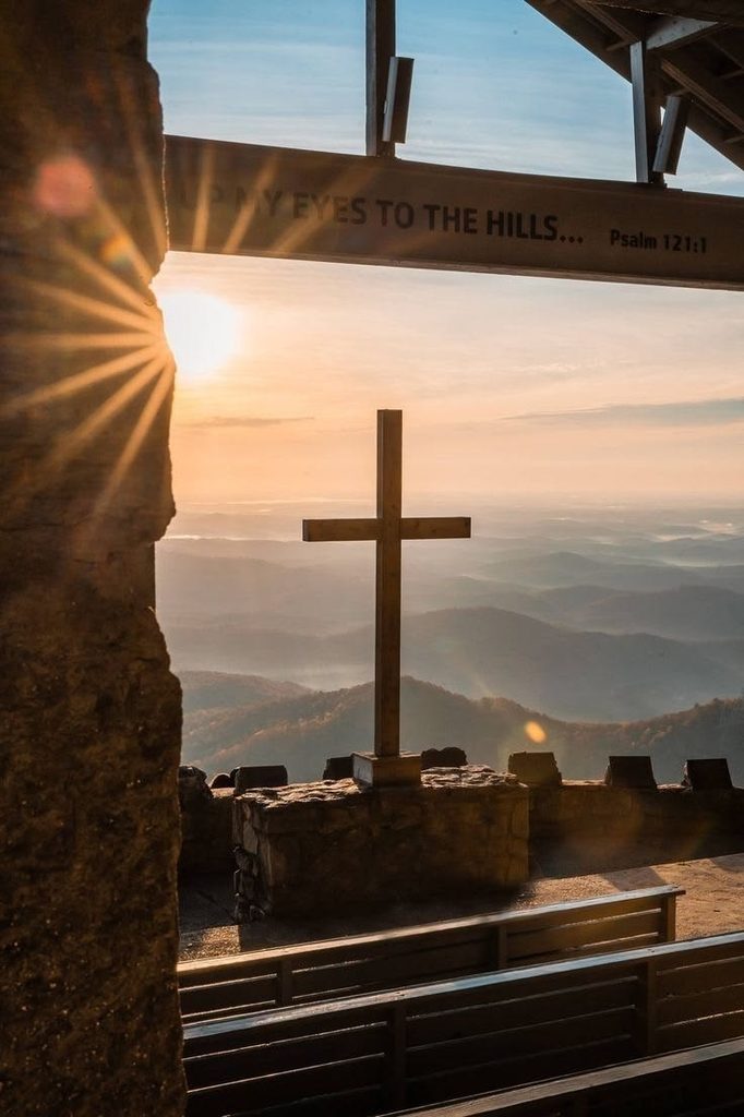 A wooden cross overlooking mountains at sunrise, framed by a stone structure with words from Psalm 121.